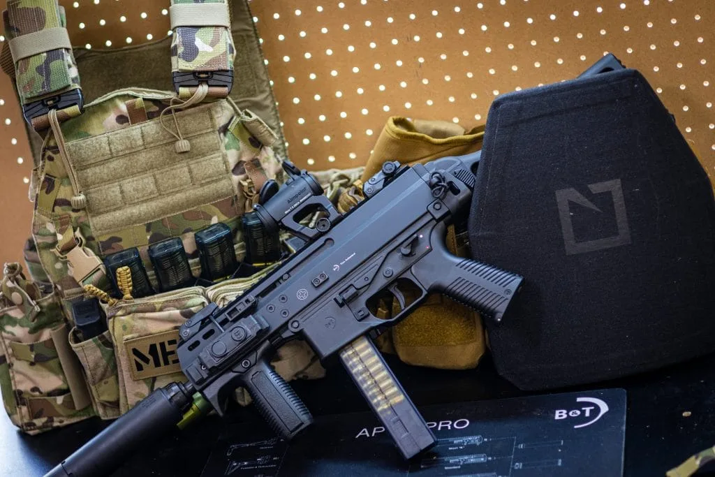 A B&T 9, plate carrier, and a HESCO L211 armor plate are placed on a surface in front of a pegboard backdrop.