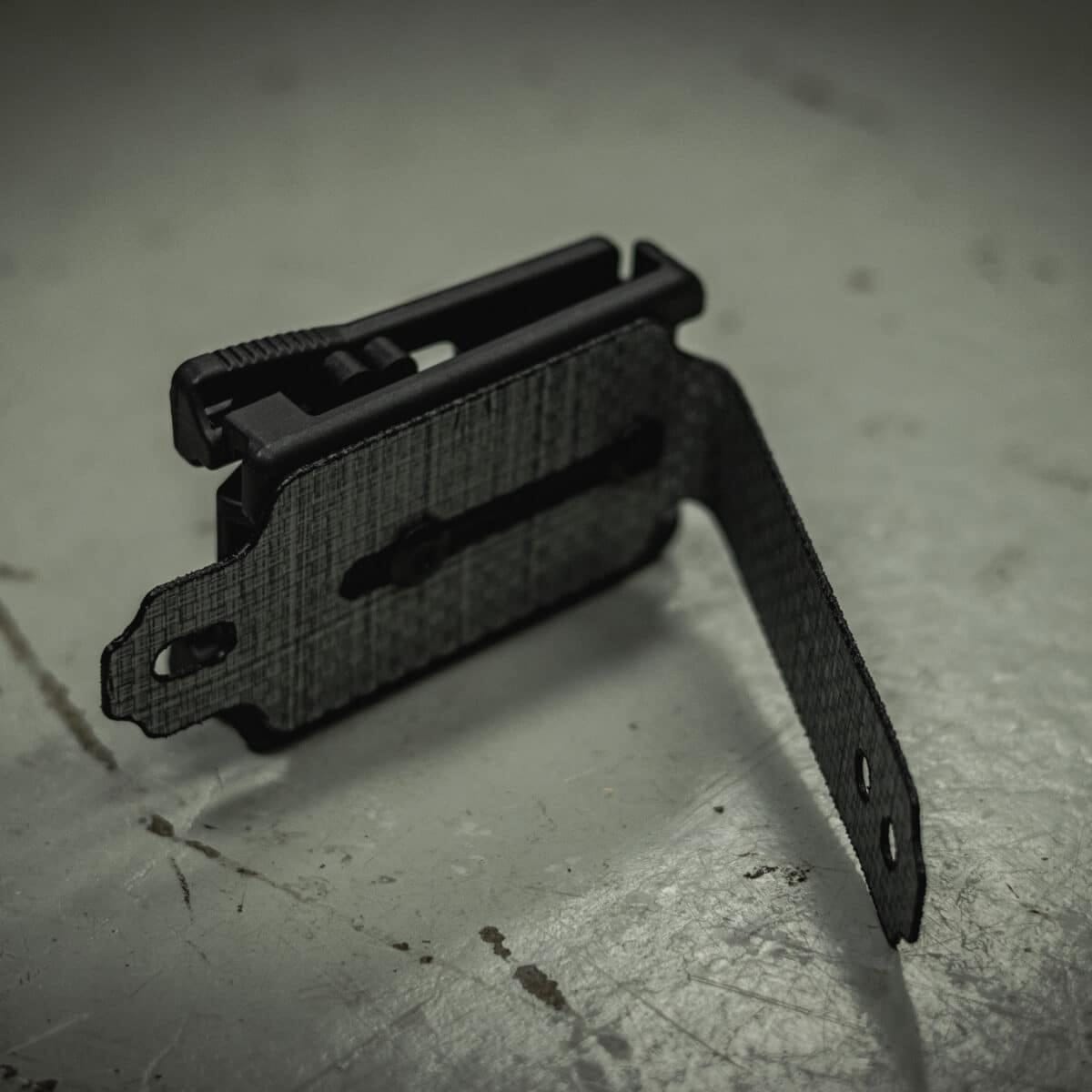 Black L-shaped TEK LOK MOLLE Adapter on grey surface.